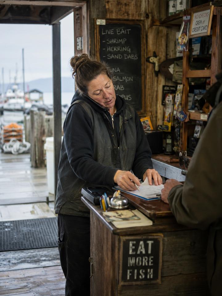 A coastal bait-shop owner taking a customer's order by hand at the counter, the harbor visible behind her