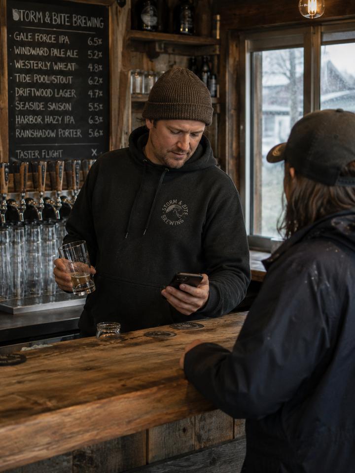 A coastal brewery owner glancing at his phone while serving a customer at the taproom counter